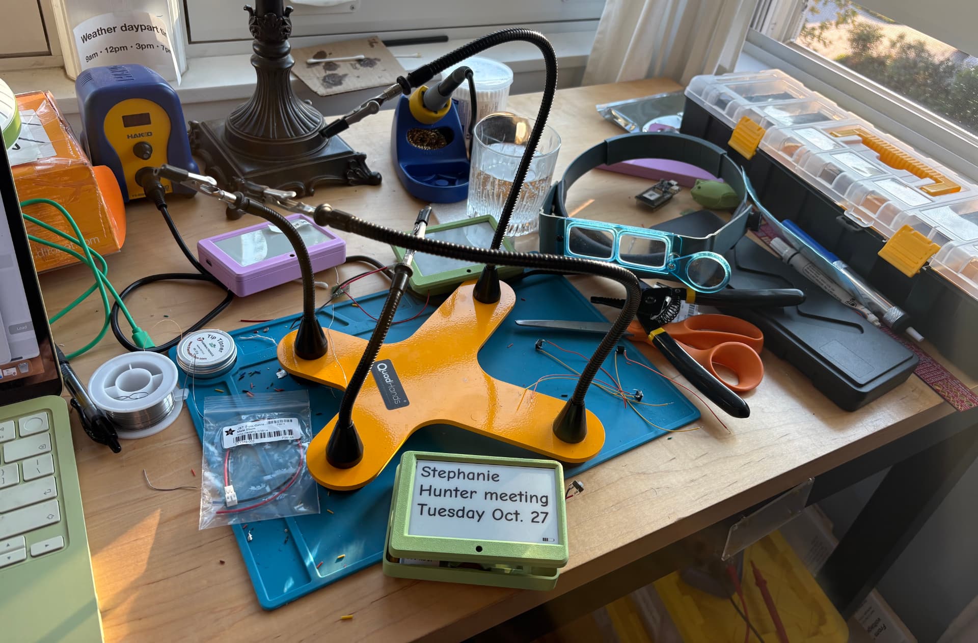 The workbench where Maggie is being built by hand in Chicago: a sage-green prototype case with a reminder on its e-ink screen, the lavender shell waiting beside it, and soldering tools spread across a blue mat.